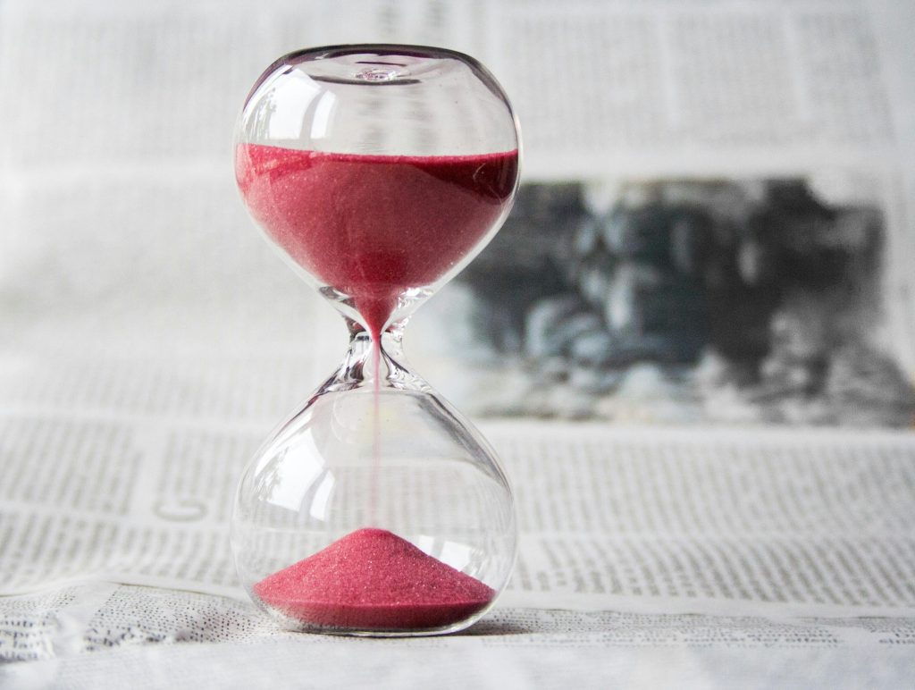 hourglass-time-hours-sand-39396-39396 Close-up of a transparent hourglass with pink sand flowing, placed on a newspaper background.