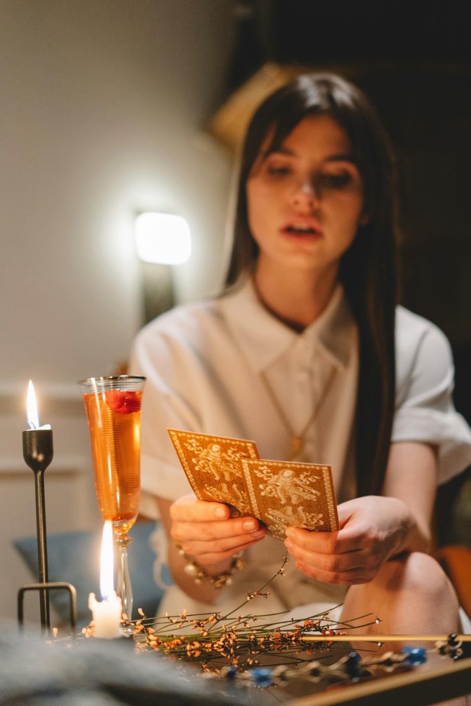 A woman reads tarot cards in a dimly lit room, with candles and a cocktail beside her, creating a mystical atmosphere.